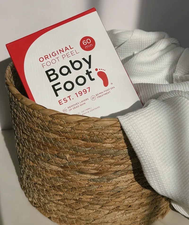 A Baby Foot Original Foot Peel box in a woven basket with a white towel