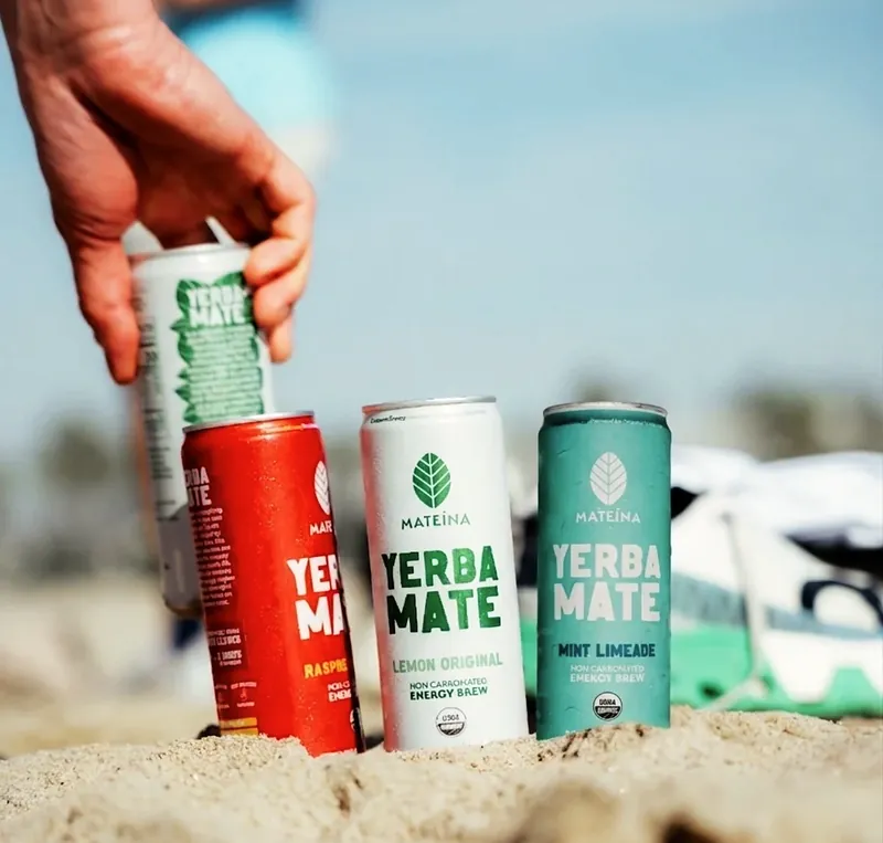 A hand placing Mateina yerba mate cans in beach sand